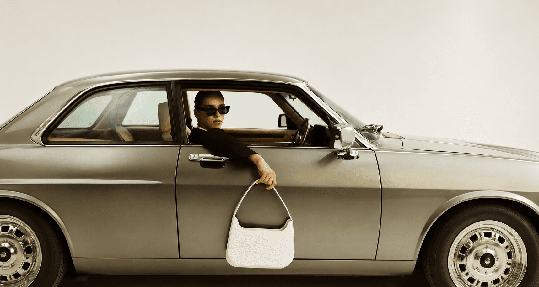 Woman in sunglasses holding an ivory handbag from the window of a silver classic car